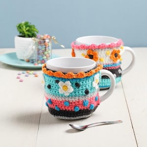 Könnte beinhalten: Zwei weiße Keramikbecher mit farbenfrohen, gehäkelten Hüllen. Die Hüllen weisen Blumen- und geometrische Muster in Blau-, Rosa-, Orange- und Schwarztönen auf. Ein kleiner Löffel liegt vor den Tassen auf dem Tisch. Eine kleine Pflanze und ein Glas Bonbons sind im Hintergrund.