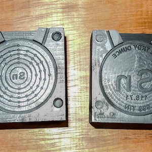 Może przedstawiać: Dwa szare metalowe formy do odlewania okrągłego monety. Forma po lewej stronie ma okrągły wzór z literą "Sn" w środku. Forma po prawej stronie ma tekst "500 ONE TROY OUNCE" i "Sn" w środku.