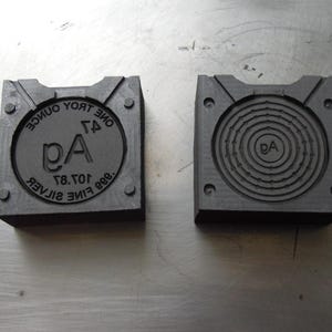 May include: Two dark gray metal molds. One mold has the text "ONE TROY OUNCE .999 FINE SILVER" around a central design with the letters "DA". The other mold has a spiral design with the letters "DA" in the center.