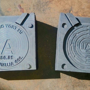 May include: Two gray metal molds with square bases. One mold has the text "13 TROY OUNCE" and "IA" with numbers and the text "MUKIMUJA COG." The other mold has concentric circles and the letters "IA".