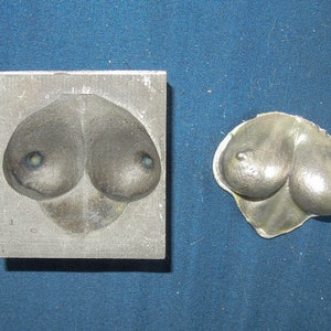 Może przedstawiać: Metalowa forma i odlew metalowy pary piersi. Forma jest kwadratowa z wypukłym wzorem dwóch piersi. Odlew jest bardziej zaokrąglony z podobnym wzorem.