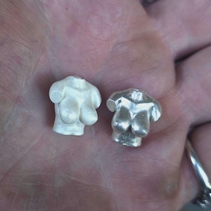 May include: Two small, three-dimensional torso charms shaped like a female bust. One charm has a matte, light-colored finish, while the other has a polished, metallic silver finish. Both pieces feature a small opening at the neck area.