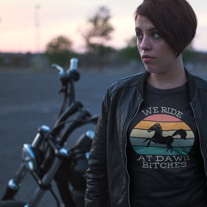May include: A woman wearing a black leather jacket and a black t-shirt with a vintage-style graphic that says "We Ride At Dawn Bitches" and a silhouette of a horse. She is standing next to a black motorcycle.