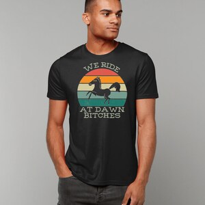 May include: Black t-shirt with a vintage-style graphic of a black horse running in front of a sunset. The graphic says "We Ride At Dawn Bitches".