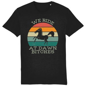 May include: Black t-shirt with a vintage-style sunset graphic and the text "WE RIDE AT DAWN BITCHES". A black silhouette of a horse is in the center of the graphic.