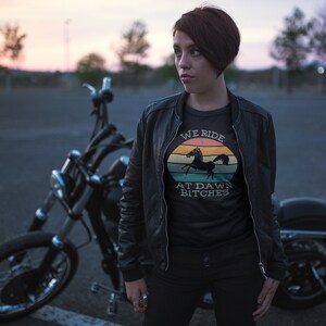 May include: A woman wearing a black leather jacket and a black t-shirt with a vintage-style graphic that says "We Ride At Dawn Bitches" and a silhouette of a horse. She is standing next to a black motorcycle.