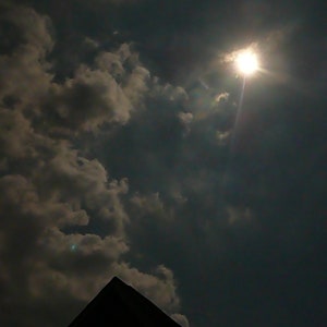 May include: A bright sun shines through dark clouds in the sky.