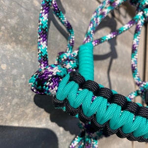 Può includere: Primo piano di un'imbracatura per cavalli con un design a corda intrecciata turchese, nera, viola e bianca. L'imbracatura presenta un capezzone turchese e una corda intrecciata. La corda è realizzata in materiale resistente.