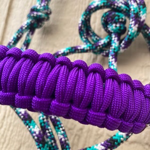 May include: Close-up of a purple and multi-colored horse halter. The halter features a braided purple noseband and coordinating rope in teal, white, black, and purple. The halter is designed for equine use.