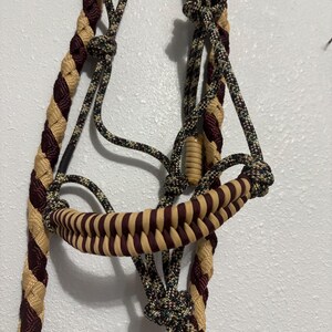 Puede incluir: Un cabestro para caballos hecho de cuerda trenzada en tonos beige, burdeos y negro. El cabestro presenta una muserola tejida y una serie de nudos y bucles para ajuste y fijación. La cuerda es gruesa y duradera.