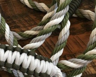 May include: A braided dog leash featuring a combination of white, gray, and olive green rope. The handle is made of tightly woven white and olive green paracord. The leash is designed for durability and style.