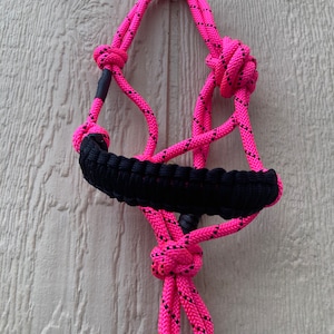 May include: A pink and black rope halter for horses. The halter is made of braided rope with black accents. The halter is hanging on a wooden wall.