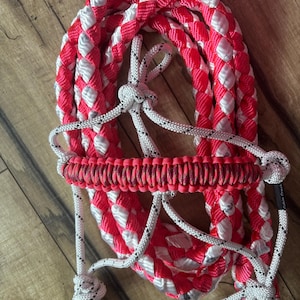 Puede incluir: Un cabezada de cuerda trenzada rosa y blanca con una muserola trenzada negra y rosa. La cabezada está hecha de cuerda gruesa y resistente y tiene un nudo seguro en la parte superior.