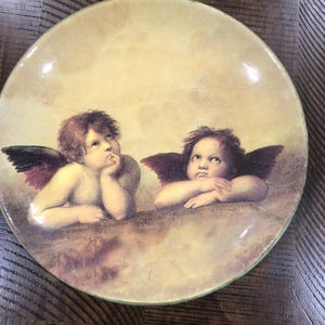 Peut inclure: Assiette décorative ronde représentant une peinture classique de deux chérubins ailés. Les chérubins ont la peau claire, les cheveux bruns et se détachent sur un fond nuageux et doux. L'assiette a un bord jaune clair et une finition brillante.