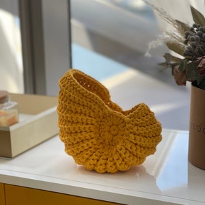 May include: A yellow crocheted basket shaped like a seashell. The basket is sitting on a white surface.