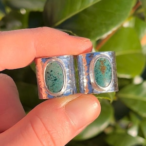 Puede incluir: Un anillo de plata con dos piedras turquesas de forma ovalada. El anillo tiene una textura martillada y se sostiene entre los dedos sobre un fondo de hojas verdes. Las piedras turquesas tienen un aspecto moteado.
