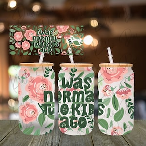 May include: Three white tumblers with a floral pattern and a bamboo lid. The tumblers have straws and the middle tumbler has the text "I was normal two kids ago" in green letters.