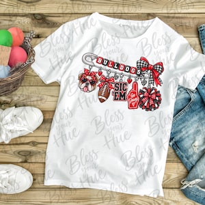May include: A white t-shirt featuring a graphic of a large safety pin with the text BULLDOGS. Hanging from the pin are charms including a bulldog wearing sunglasses, a football, a red foam finger with GO DAWGS, a pom-pom, and plaid bows.