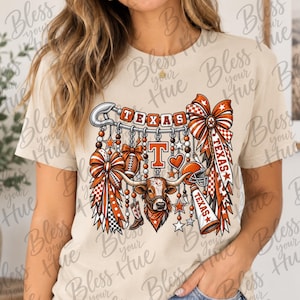 May include: A cream-coloured t-shirt featuring a graphic of a large safety pin adorned with orange and white charms, including a longhorn steer, a football, a megaphone with the text Texas, a cowboy boot, stars, hearts, and bows with Texas text.