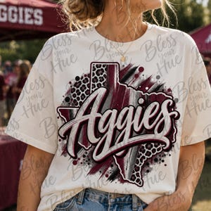 Puede incluir: Una camiseta de manga corta color crema con un gráfico del estado de Texas en granate, negro y blanco. El diseño incluye un estampado de leopardo, texturas de pinceladas y la palabra Aggies escrita en una fuente cursiva blanca grande.