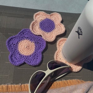 May include: Three crocheted flower-shaped coasters in pink and purple, with one coaster supporting a white water bottle and a pair of sunglasses resting on top of the coasters.