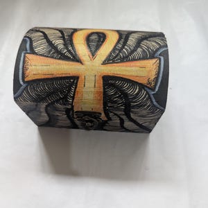 May include: A small, black wooden box featuring an Ankh symbol painted in gold and orange on the lid. The Ankh is set against a detailed, swirling black and white pattern. The box is secured with a small metal clasp.