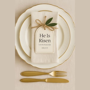 May include: A formal place setting featuring a white plate with gold trim, and a folded white napkin. A tag with the text "He Is Risen" is tied to the napkin with twine and olive branches. A gold fork and knife are placed below.