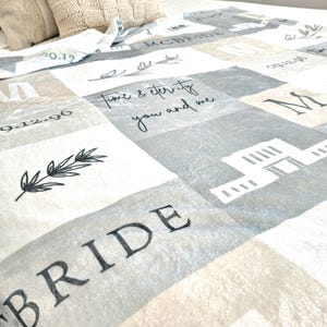 May include: A white, gray, and beige fleece blanket with a checkered pattern. The blanket has the words "Bride" and "Time & Eternity You and Me" printed on it. The blanket also has the dates "09.12.96" and "20.12" printed on it.