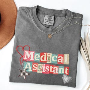 May include: A gray t-shirt with the words "Medical Assistant" in colorful block letters. A red stethoscope, a black and white star, and a silver necklace with a pendant are on the shirt. The shirt is folded on a white surface.