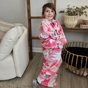 Op de afbeelding: Een jong persoon gewikkeld in een roze en witte deken met aardbeienpatroon. De deken heeft een herhalend ontwerp van aardbeien en tekst. De persoon glimlacht en staat in een kamer met een houten ladder en een witte stoel.