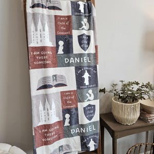 May include: A white fleece blanket with a colorful patchwork design featuring images of a temple, an open book, and a child praying. The blanket has the text "I am a Child of the Covenant", "I am a Child of God", "I am a Disciple of Jesus Christ", and "Daniel" repeated throughout the design.