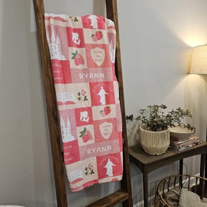 Op de afbeelding: Een rood-wit patroon deken met diverse afbeeldingen, waaronder aardbeien, een kerk en de tekst "Choose the Right" en "RYANN". De deken is over een houten ladder gedrapeerd.