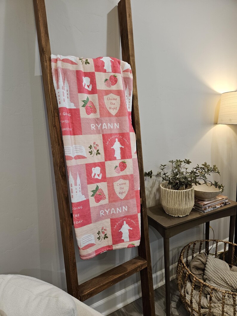 Op de afbeelding: Een rood-wit patroon deken met diverse afbeeldingen, waaronder aardbeien, een kerk en de tekst "Choose the Right" en "RYANN". De deken is over een houten ladder gedrapeerd.