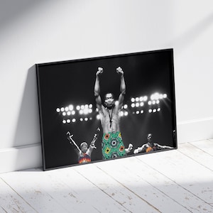 May include: Black and white framed print of a man with his arms raised in a concert setting. The man is wearing a patterned wrap. Stage lights illuminate the background. The print is leaning against a white wall.