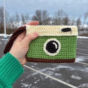 Könnte beinhalten: Eine handgefertigte Kameratasche aus Häkelarbeit in Grün, Creme und Braun. Die Tasche hat ein schwarz-weißes kreisförmiges Design, einen Reißverschluss und eine braune Handschlaufe. Die Tasche wird vor einem bewölkten Himmel gehalten.