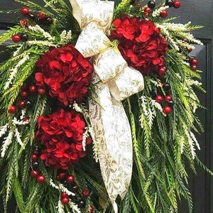 May include: A festive wreath featuring vibrant red hydrangeas, berries, and lush green foliage. A cream-colored ribbon with a gold pattern adds an elegant touch. This decorative wreath is perfect for seasonal home decor.