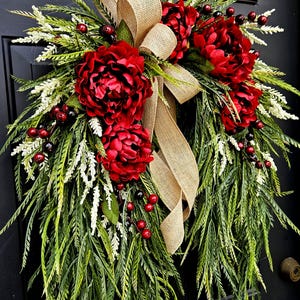 May include: A decorative wreath featuring vibrant red peonies, green foliage, white accents, and red berries. A burlap ribbon adds a rustic touch. The wreath is designed for door or wall decor.