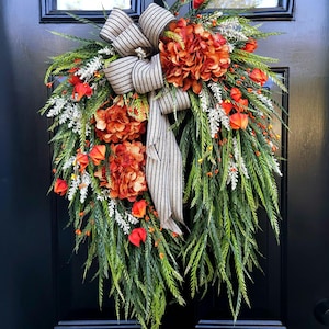 May include: A decorative fall wreath with orange and red flowers, white accents, and green foliage. A striped bow adds a rustic touch. The wreath is displayed on a black door, perfect for seasonal home decor.