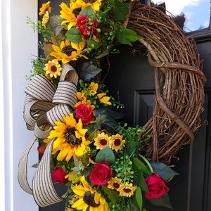 Pu&ograve; includere: Una ghirlanda decorativa con una base in rami di vite naturali marroni, decorata con girasoli giallo brillante, rose rosso scuro e fogliame verde. Un grande fiocco in tessuto a righe beige e nero &egrave; fissato lateralmente per aggiungere consistenza.