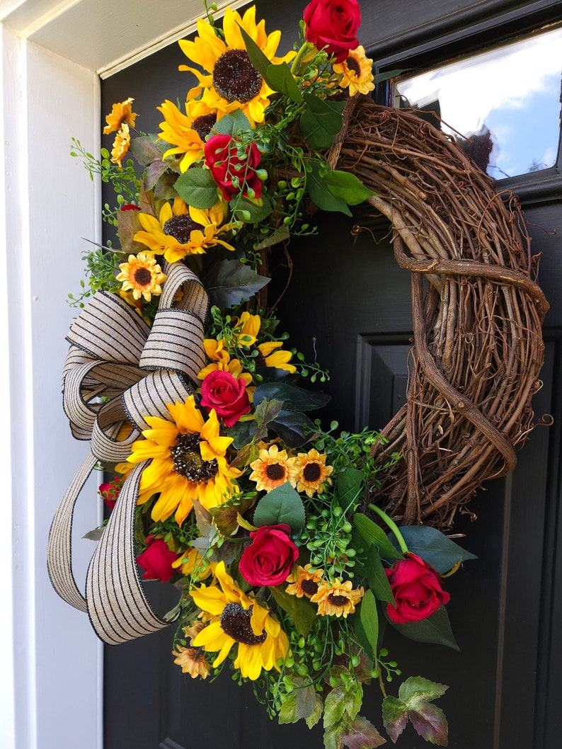 Pu&ograve; includere: Una ghirlanda decorativa con una base in rami di vite naturali marroni, decorata con girasoli giallo brillante, rose rosso scuro e fogliame verde. Un grande fiocco in tessuto a righe beige e nero &egrave; fissato lateralmente per aggiungere consistenza.