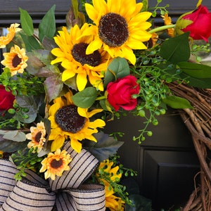 Pu&ograve; includere: Una ghirlanda decorativa con girasoli giallo brillante e rose rosso scuro disposti su una base di ramoscelli marroni naturali. La composizione floreale &egrave; arricchita da fogliame verde e da un grande fiocco in tessuto beige a righe nere.
