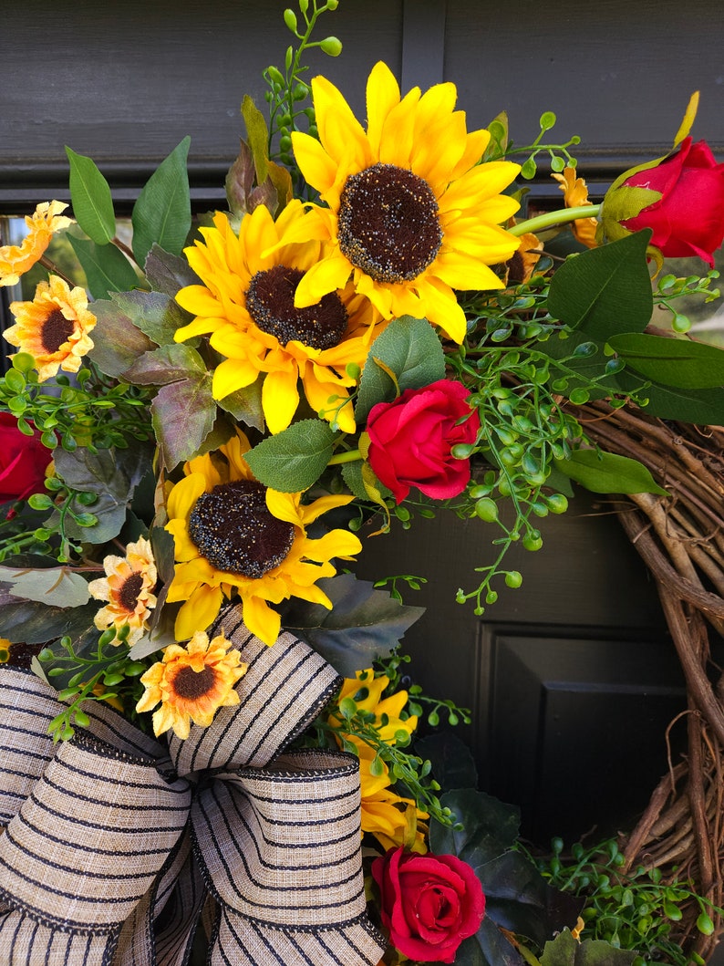 Pu&ograve; includere: Una ghirlanda decorativa con girasoli giallo brillante e rose rosso scuro disposti su una base di ramoscelli marroni naturali. La composizione floreale &egrave; arricchita da fogliame verde e da un grande fiocco in tessuto beige a righe nere.