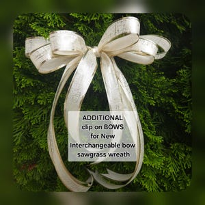 May include: A decorative white bow with gold trim, set against a backdrop of green foliage. The bow features multiple loops and long, flowing ribbons. Text on the image reads: "ADDITIONAL clip on BOWS for New Interchangeable bow sawgrass wreath."