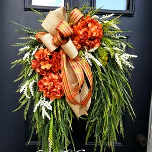 May include: A fall-themed door wreath with orange hydrangeas, green foliage, and a burlap bow with orange and yellow plaid ribbon. The word "Welcome" is at the bottom of the wreath.