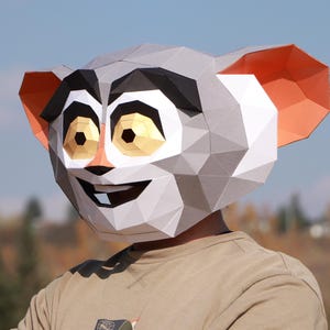 May include: A geometric paper mask of a lemur with a smiling expression. The mask features a grey, white, black, orange, and gold colour scheme. The person wearing the mask is wearing a tan shirt.