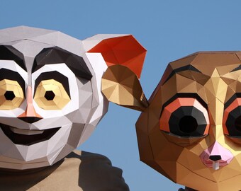 May include: Two geometric lemur masks, one gray and white with orange ears, the other brown and gold. The masks have large eyes and are worn against a clear blue sky. One person is giving a peace sign.