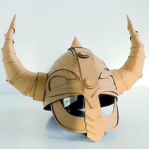 May include: A handcrafted Viking helmet made from cardboard, featuring prominent, curved horns. The helmet is constructed from corrugated cardboard, with visible seams and edges. The design includes a pointed top and a face guard, showcasing a DIY craft project.