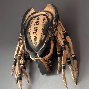 May include: A sculpted mask featuring a black and tan color scheme with a textured, weathered finish. The mask displays intricate black symbols on the forehead and includes long, dangling, rope-like appendages on both sides with black bands.