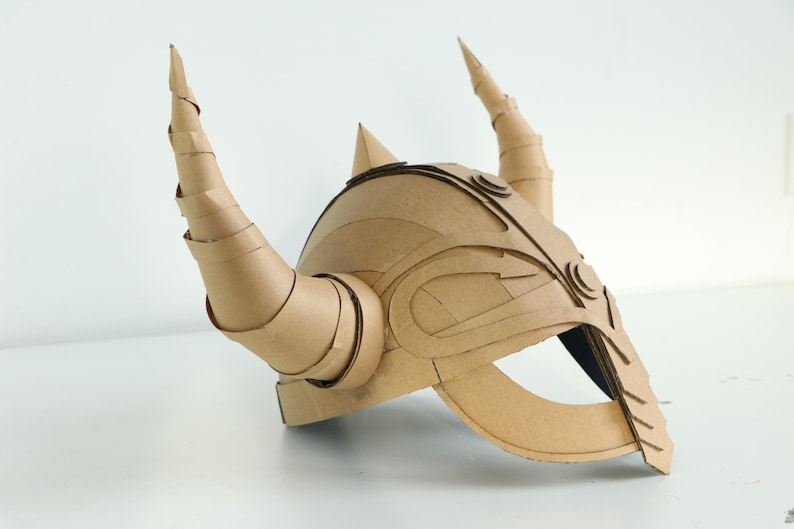 May include: A handmade cardboard helmet with large, spiraling horns and intricate layered details. The helmet is a natural tan color and appears to be constructed from multiple pieces of corrugated cardboard, suggesting a DIY or costume theme.