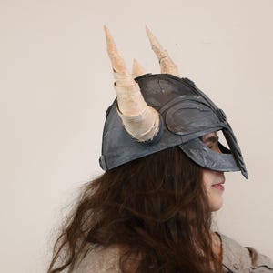 May include: A gray Viking helmet with two large, cream-colored horns. The helmet has a dark gray finish and is worn by a person with long, brown hair. The helmet is a costume piece.